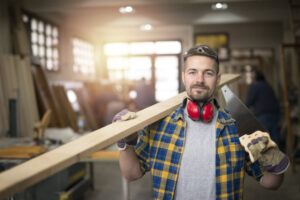 Portrait of professional middle aged carpenter with wood plank and tools standing in his woodworking workshop.